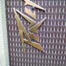 Detail kovového loga v hnědo-zlaté barvě s ostrými hranami, které je připevněno na hnědou textilní látku. Logo tvoří stylizované geometrické tvary, k…