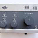 Audio rozhraní s šedým kovovým šasi a dřevěnými bočními panely. Na horní části je logo UA v kosočtverci a LED indikátory s nápisy INPUT, OUTPUT a HOS…