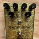 Zlatý kytarový efekt s nápisem PUGILIST DISTORTION a logem Fender. Na horní straně jsou čtyři černé otočné knoflíky s popisky TONE A, GAIN A, TONE B…