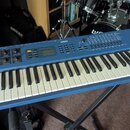 Modrý syntezátor značky YAMAHA s modelem CS1X CONTROL SYNTHESIZER. Nástroj má bílé a černé klávesy, displej a řadu ovládacích tlačítek a posuvníků. P…