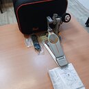 Na fotografii je stříbrný bubnový pedál s oranžovými detaily a nápisem IDemon. Vedle něj leží černý přepravní kufr s logem Pearl, náhradné části v pl…