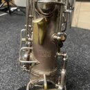 Detailní záběr na část saxofonu v matném stříbrném provedení. Na korpusu je vyryto: "Patented in USA 11082", pod tím písmeno "A" a sériové číslo "№ 2…