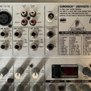 Stříbrný mixážní pult značky BEHRINGER s označením EURORACK® UB2442FX-PRO. Na horní části jsou vidět různé vstupy a výstupy, včetně konektorů pro mik…