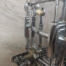 Detailní záběr na chromovaný mechanismus snare drumu. Jsou vidět kovové napínací šrouby, písty a plastové bílé vložky. Povrch chromu vykazuje známky…