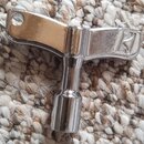 Kovový klíč v tvaru T s chromovaným povrchem. Na spodní části rukojeti je vyryto logo ve tvaru stylizovaného písmena M. Předmět leží na textilním kob…
