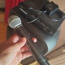 Černý bezdrátový mikrofon s kovovou mřížkou, na kterém je nápis THE SHURE WIRELESS. V pozadí je černý přijímač s nápisem SHURE a nadpisem SCH RECEIVE…