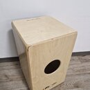 Dřevěný cajon světlé barvy s přirozenou strukturou dřeva. Na horní desce je nápis „Meal Autumn Edition“ a v dolní části přední stěny se nachází logo…