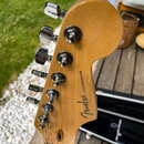 Záběr na zadní stranu hlavy kytary z přírodního dřeva s chromovanými ladičkami. Na dřevě je černý nápis Fender STRATOCASTER. Povrch dřeva a chromové…