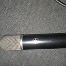 Černý kondenzátorový mikrofon s kovovou síťovkou v horní části. Na těle je umístěno kulaté logo s nápisem AKG. Povrch mikrofonu vykazuje drobné odpad…
