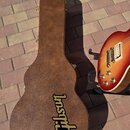 Hnědý kytarový kufr z texturovaného materiálu s žlutým logem Gibson. Vedle kufrů je vidět část elektrické kytary s tělem v barvě sunburst, dvěma humb…