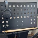 Černý kovový držák na příslušenství s řadou kulatých otvorů. Na držáku visí černá sluchátka s bílým nápisem Roland RH-5. V dolní části na polici leží…