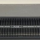 Černý rackový zesilovač s nápisy QSC POWERLIGHT 2, 1800 WATT PROFESSIONAL AMPLIFIER a PL218 CUSTOM ENGINEERED FOR TOURING. Na pravé straně jsou dva o…
