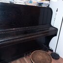 Černý piano v provedení s viditelnou strukturou dřeva. Na horní desce leží stoh papírů a notového zápisu. Před nástrojem stojí kulatý dřevěný taburet…