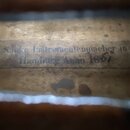 Detail vnitřku hudebního nástroje s nalepeným papírovým štítkem. Na štítku je čitelný nápis: „I. Suke Instrumentenmacher in Hamburg Anno 1857“. Papír…