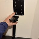 Černý kovový držák na nástroj připevněný ke stěně. Sestává z ploché základní desky s výřezy a kolmého ramene s vložným prvkem. Na povrchu nejsou vidi…
