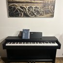 Černé digitální piano s bílými klávesami a třemi pedály. Na horní části je viditelné logo YAMAHA. Na nástroji leží modrá kniha s nápisy YAMAHA, ARIUS…