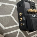 Černý elektronický přístroj s nápisem BEATB. Na boku jsou umístěny tři zlaté konektory s popisky R, L (Mono) pod společným označením Output a Phones…