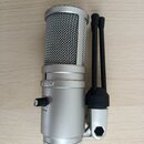 Stříbrný kondenzátorový mikrofon s kovovou mřížkou a černým stativem. Na těle mikrofonu je viditelné logo Redragon a nápis VOLUME u ovládacího knoflí…