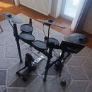 Elektronická bicí souprava v černé barvě s gumovými pady a plastickými činely. Na ovládacím modulu je viditelné logo Roland. Souprava obsahuje pedály…