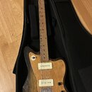 Elektrická kytara s přírodním dřevěným povrchem těla a hnedým hmatníkem, umístěná v černém obalu. Na hlavě je viditelné logo "Fender". Kytara má dva…