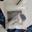 Bílý plastový zip-lock sáček, ve kterém se nachází bílá látka a tmavý předmět. V pozadí je vidět část bílého kytarového mostu s bílými knoflíky a nap…