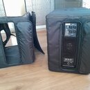 Dvě černé transportní tašky z látkového materiálu. Pravá taška obsahuje reproduktor, na jeho zadním panelu je viditelný nápis SUB 702-AS a ovládací p…