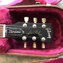 Hlava elektrické kytary v černé barvě, položená v otevřeném kufru s fialovým polstrováním. Na hlavě jsou bílé nápisy „Gibson“, „Les Paul MODEL“ a v r…