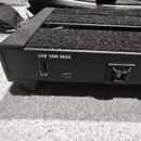Detail černé kovové konstrukce s černým textilním povrchem. Na boku jsou umístěny USB porty a napájecí konektor s nápisem USB 18W MAX. Povrch je matn…