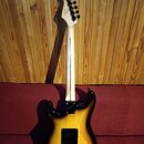 Zadní strana elektrické kytary s tělem v žlutém gradientu (sunburst) a světlým javorovým hnedkem. Na hlavě je viditelné logo Fender. Kytara má černé…
