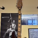 Elektrická basová kytara s tmavě hnědým tělem a světle dřevěným hmatníkem. Na hlavě nástroje je nápis Squier PRECISION BASS. Kytara visí na stěně ved…