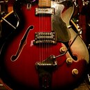 Polodupatá elektrická kytara s červeným gradientním povrchem a černým rýhovaným plecháčem. Nástroj má dva humbuckery, tři ovládací knoflíky a vibračn…