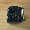 Černý elektronický přístroj s kovovou rukojetí na pravé straně. Na zadní straně jsou dva velké konektory, nápisy CAMERA IN, REMOTE a CAMERA OUT, přep…