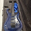 Modrá elektrická kytara s plamenovitým vzorem dřeva v černém tvrdém kufru. Na hlavě hрифu je bílý nápis "Cort". Kytara má tři humbuckery, černý most…