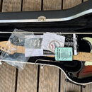 Elektrická kytara s tělem v provedení sunburst a bílým pickguardem leží v černém tvrdém kufru. Na hlavě kytary je viditelné logo Fender a nápis STRAT…