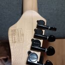 Zadní strana hlavy světlé dřevěné kytary s pěti černými ladicími mechanismy. Na mechanismech je vyryté logo SCHECTER. Na dřevě jsou nápisy Complies w…