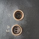 Detail dvou kulatých konektorů zasazených do černého povrchu s texturou kůže. Horní konektor má vnitřní piny, vedle něj je nečitelný symbol. Dolní ko…