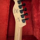 Záběr na hřif a hlavu světle dřevěné kytary s šesti černými ladičkami. Na zadní straně hlavy je nápis „Designed and Based by Fender“, text „Crafted i…