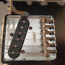 Detail elektrické kytary s černým snímačem a kovovým mostkem. Na kovové části je vyryt nápis FENDER. Povrch kovu vykazuje známky opotřebení a drobné…
