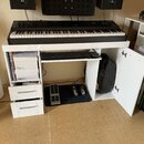 Na bílém stole stojí černý digitální klavír s bílými klávesami. Pod stolem jsou umístěny dvě sustain pedály, z nichž jedna má nápis ROLAND. Vlevo jso…