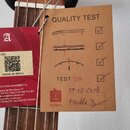 Detail krku kytary s dvěma přivázanými štítky. Červený štítek obsahuje logo "A", nápis "7800 Z-NATURE IGOG7C", QR kód, text "MADE IN SPAIN" a kontakt…