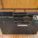 Zadní strana černého elektronického zařízení s kovovým šasi a ventilační mřížkou. Vlevo jsou čtyři konektory, vpravo je vidět bílý napájecí kabel. Up…