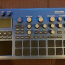 Modrý sekvenční syntetizátor s nápisy electribe MUSIC PRODUCTION STATION a KORG. Zařízení obsahuje displej, několik otočných knoflíků rozdělených do…