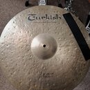 Kulatý mosazný činel s kovaným povrchem. Na horní části je černý nápis Turkish a pod ním text handmade cymbals from Turkey. Ve spodní části je rukopi…