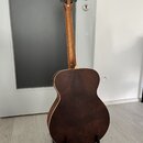 Akustická kytara v tmavě hnědé barvě s viditnou strukturou dřeva. Na hlavě nástroje je bílý nápis "Taylor". Kytara stojí ve svislé poloze na černém s…