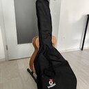 Akustická kytara v dřevěném provedení umístěná na stojanu, přes kterou je navlečen černý textilní obal. Na hlavě kytary je bílý nápis „Cort“. Na spod…