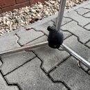 Detail spodní části kovového stativu s chromovaným povrchem. Stativ má tři rozkládací nohy spojené černým plastovým kloubem s úchytem. Povrch kovovýc…
