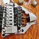 Detail chromovaného tremolo systému na kytáře s výraznou dřevěnou strukturou povrchu. Na kovovém dílu jsou viditelné nápisy „Floyd Rose“ a „FRX“. Sys…