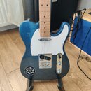 Modrá elektrická kytara typu Telecaster s bílým pickguardem a světlou hnedou hmatníkovou deskou. Na spodní části těla je vlevo umístěná kulatá černob…