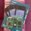Fotografie zobrazuje červenobílý karton s nápisem YAMAHA PLG150-AN Plug-in Board, modrou obálku s logem YAMAHA a čtyři 3,5" diskety. Na disketách je…