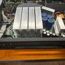 Fotografie zachycuje vnitřek otevřeného elektronického zařízení, pravděpodobně zesilovače. Uvnitř jsou vidět velké hliníkové chladiče, modré kondenzá…